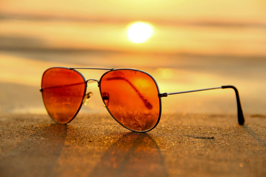 A pair of orange-tinted aviator sunglasses rests on wet beach sand, with the sun setting over the water in the background.