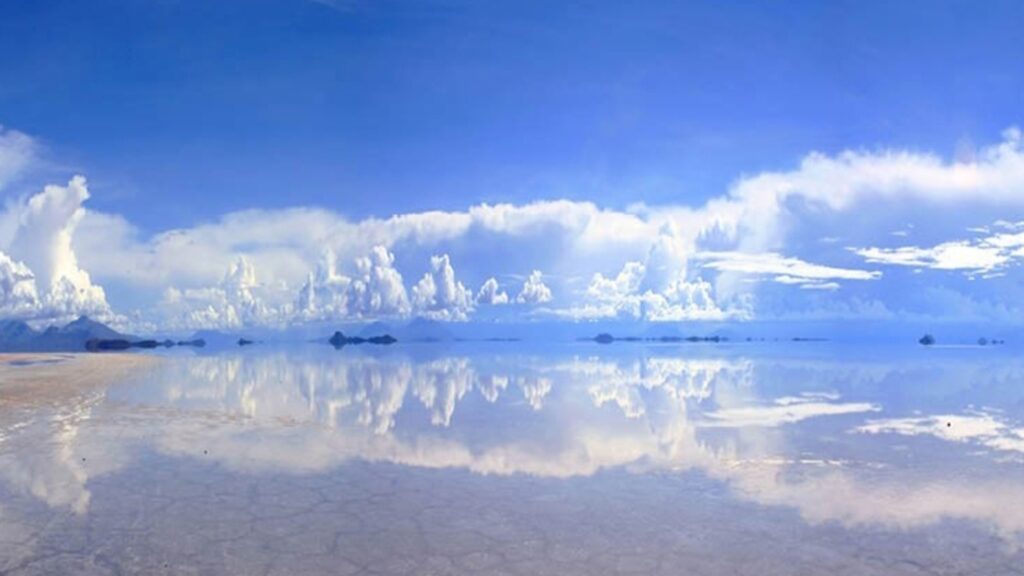 A vivid blue sky with towering white clouds reflected across a shallow, mirror-like salt flat, with low dark hills on the horizon.