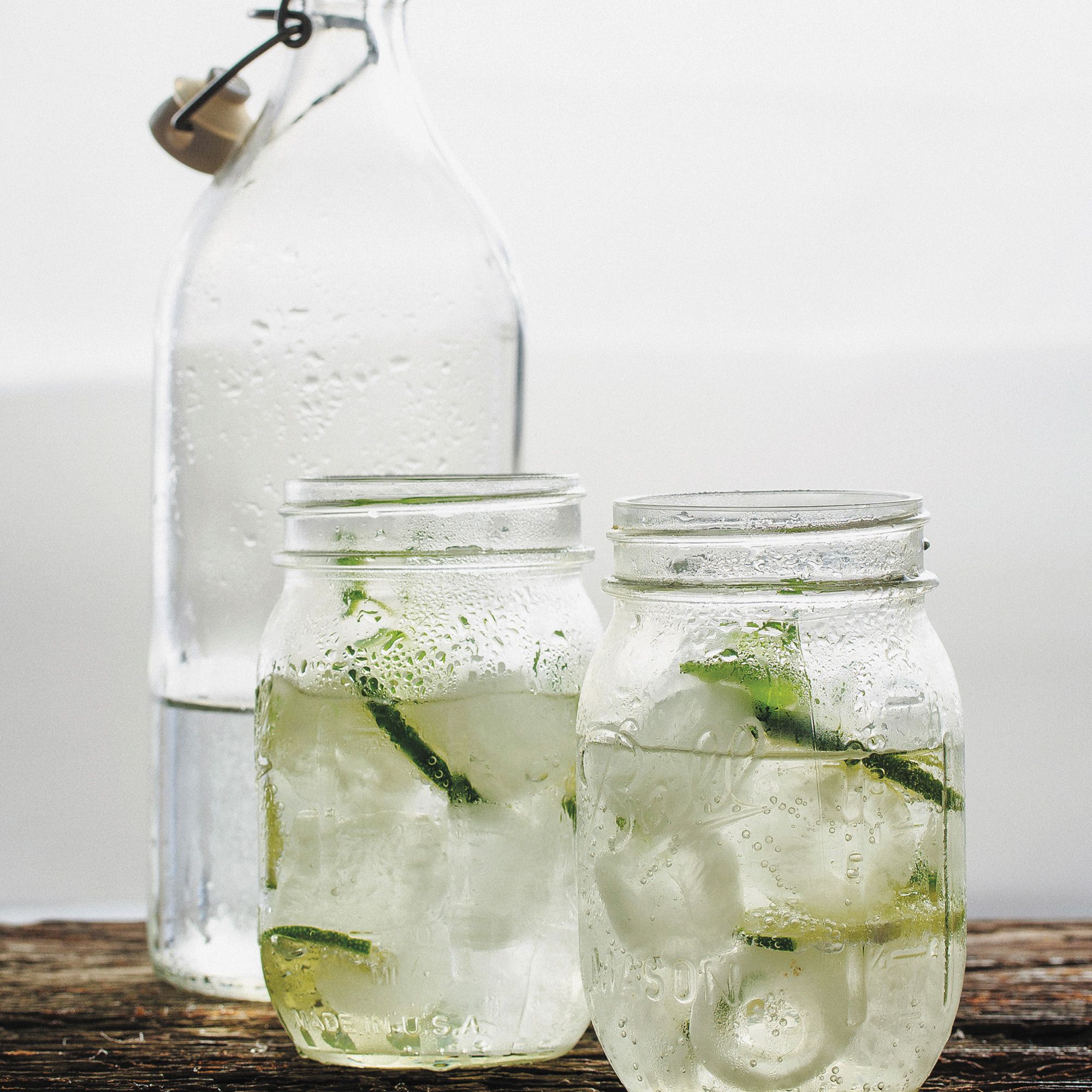 Glass jars of a pale drink with ice and lime slices sit in front of a clear bottle.