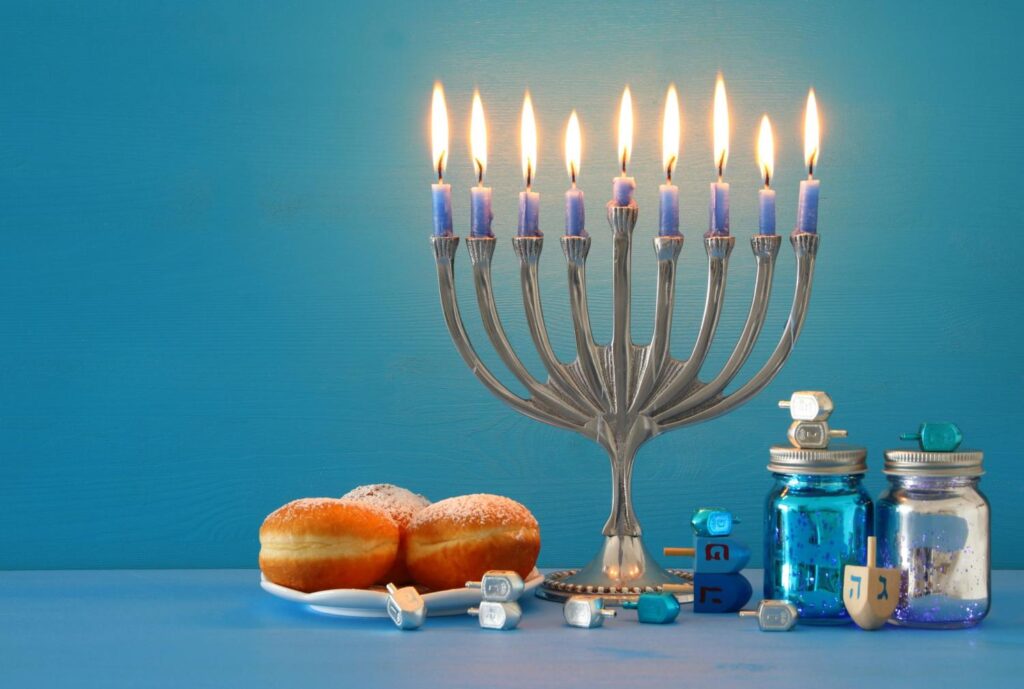 A silver Hanukkah menorah with nine lit candles stands beside sufganiyot doughnuts and blue jars and dreidel decorations.