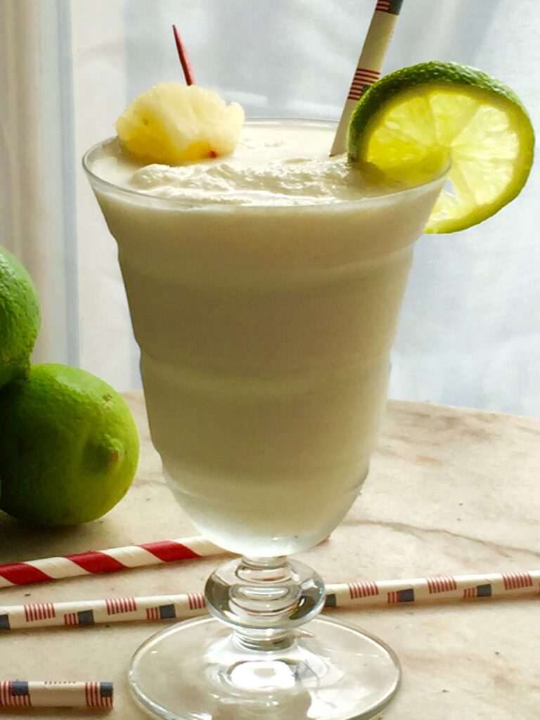 A creamy pale drink in a stemmed glass, garnished with a lime wheel and served beside whole limes and striped straws.