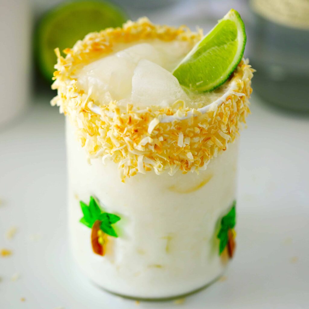 A creamy white drink in a glass, rimmed with toasted coconut and topped with ice and a fresh lime wedge.