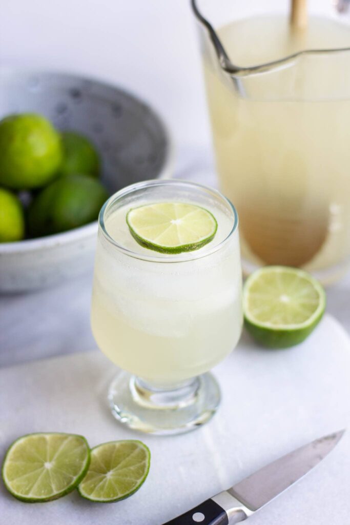 A glass of pale lime drink garnished with a lime slice, with a pitcher and fresh limes nearby.
