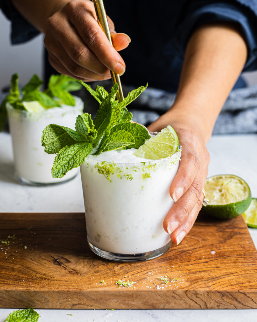 A creamy white drink topped with mint leaves, lime, and coarse salt is being stirred in a glass, with another glass and cut lime nearby.