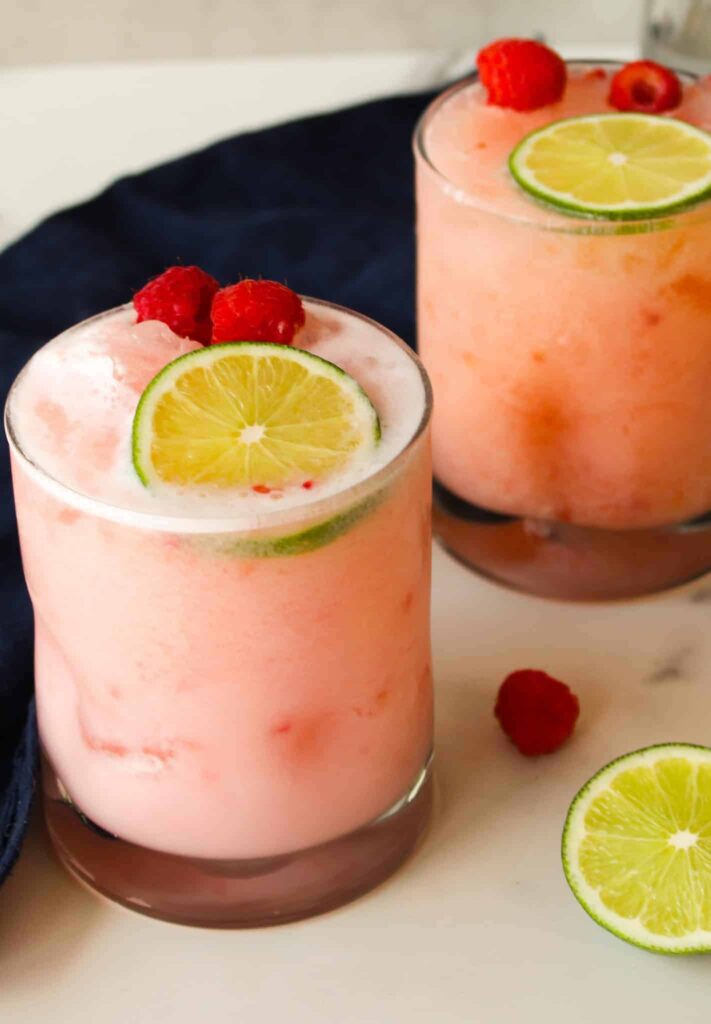 Two glasses of frothy pink drink garnished with lime slices and raspberries, with extra lime and raspberries on the table.