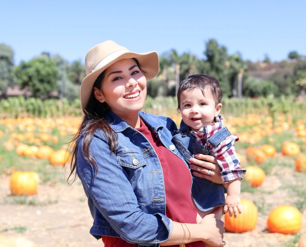 A smiling woman in a sun hat holds a baby among pumpkins in a sunny field.