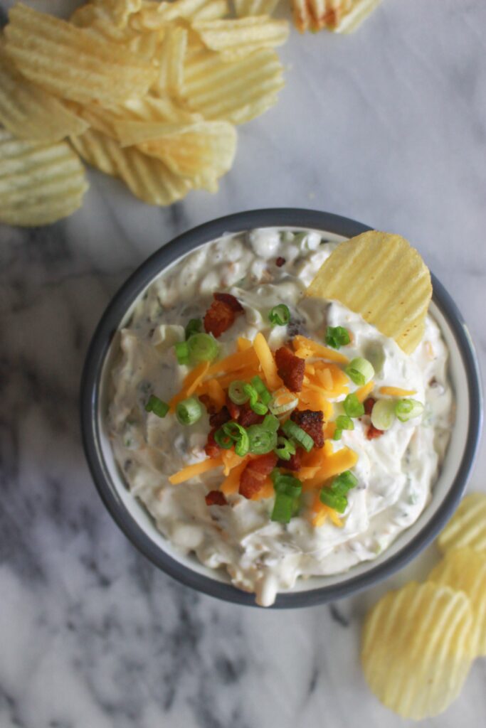 A bowl of creamy dip topped with shredded cheese, bacon pieces, and sliced green onions, surrounded by ridged potato chips.