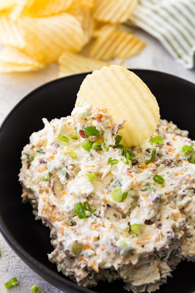 A creamy, chunky dip topped with sliced green onions sits in a black bowl, with a ridged potato chip nestled in the dip and more chips in the background.