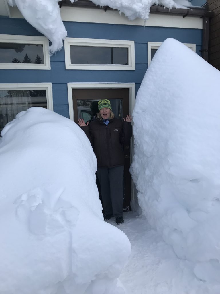 A person in a winter hat stands in a doorway between towering snowdrifts beside a blue house, with heavy snow piled on the roof.