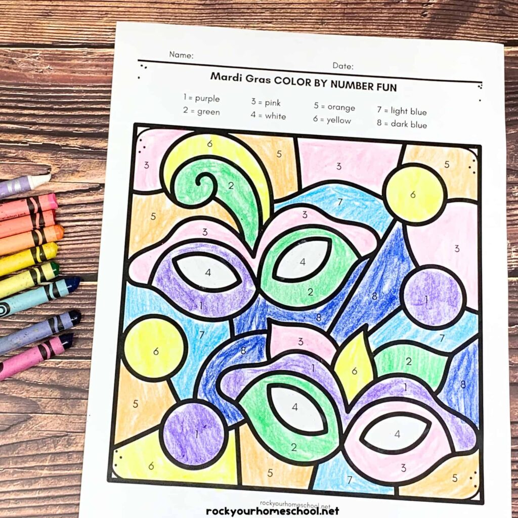 A completed Mardi Gras color-by-number page with colorful masks, beads, and numbered sections, alongside crayons on a wooden surface.