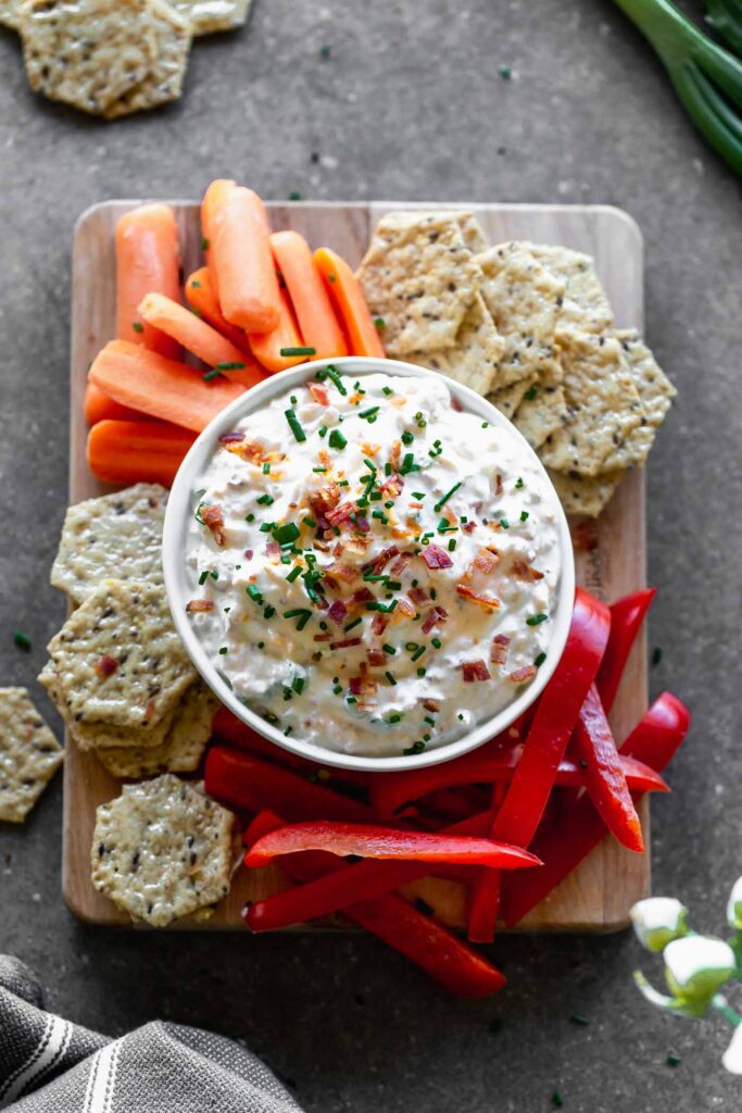 A bowl of creamy dip topped with chopped chives and crisp bacon pieces, surrounded by seeded crackers, carrot sticks, and red pepper strips.