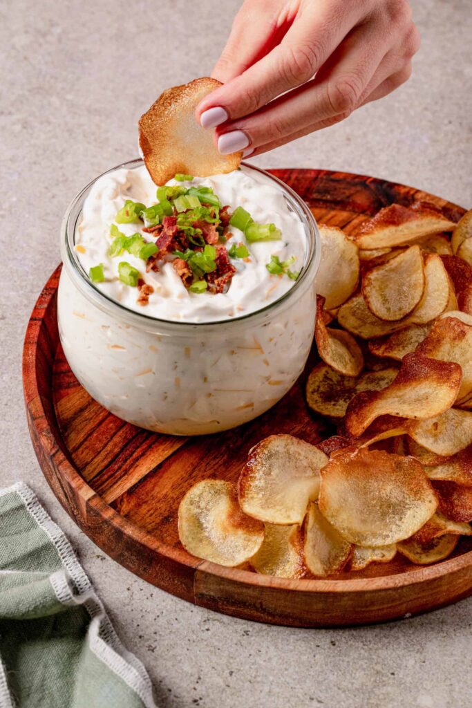 A hand dips a potato chip into a bowl of creamy dip topped with bacon bits and chopped green onions, surrounded by chips on a wooden platter.