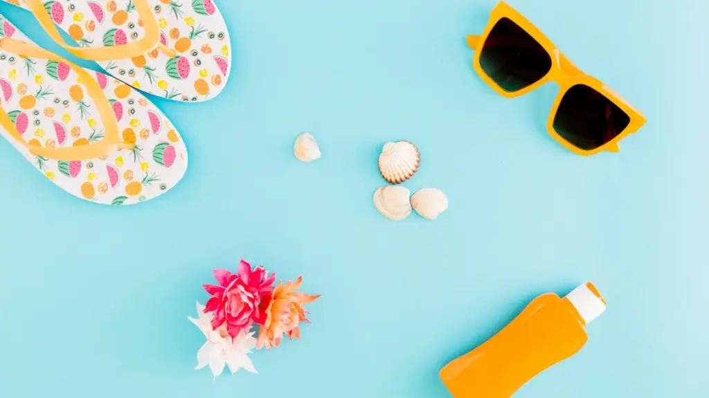 Flat lay of summer accessories on a turquoise background: fruit-patterned flip-flops, yellow sunglasses, seashells, bright flowers, and a bottle of sunscreen.