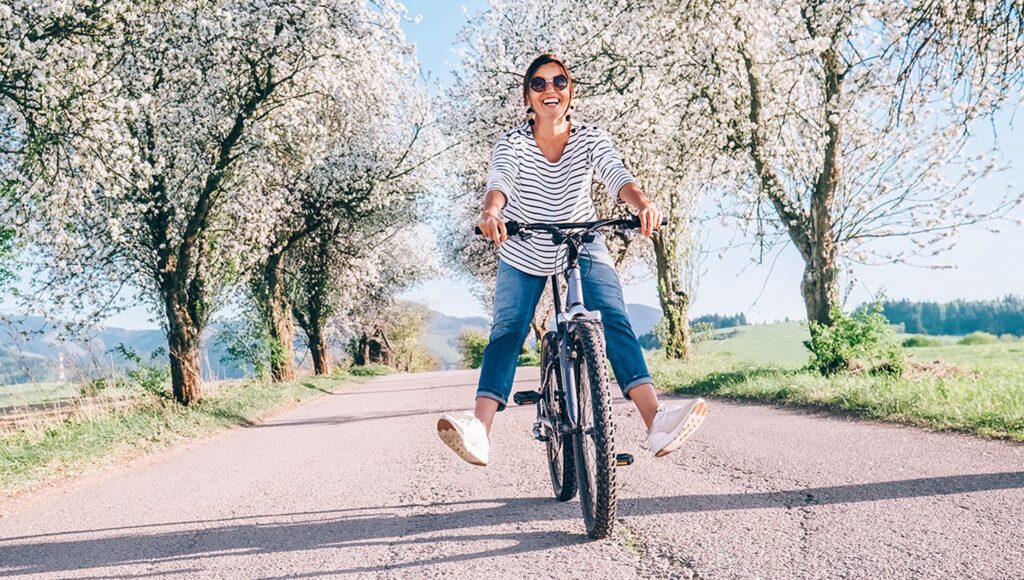 A woman rides a bicycle along a country road beneath flowering trees on a bright spring day.