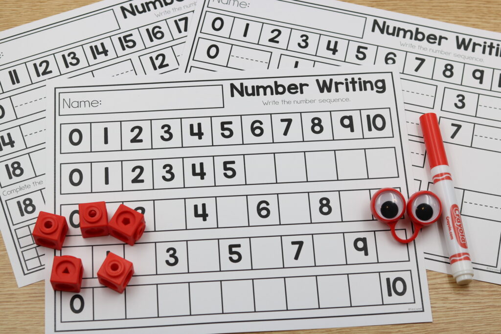 Several number-writing worksheets show sequences from 0 to 10 with blank boxes for students to complete. Red manipulatives and a marker lie on the pages.