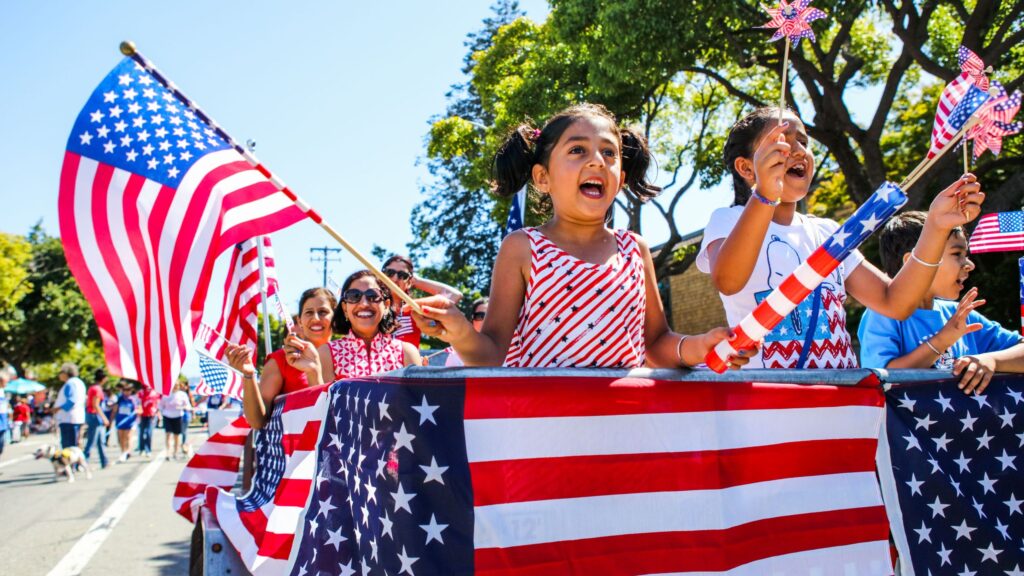 Children celebrate outdoors behind American flags, waving patriotic pinwheels at a parade.