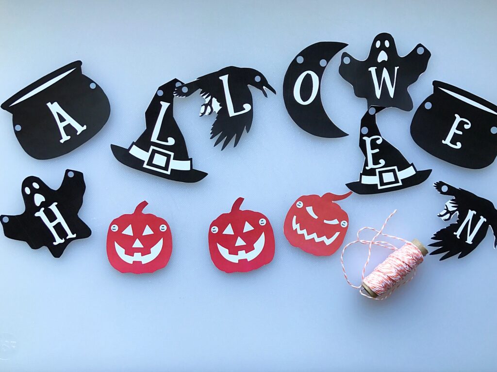 Paper Halloween banner pieces with black letters, ghost and witch-hat shapes, a raven, and three red jack-o’-lanterns are arranged on a light surface beside a spool of twine.