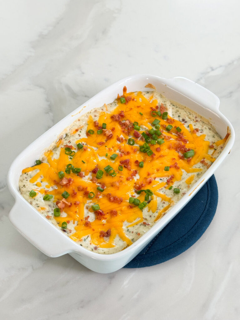 A creamy dip topped with melted cheddar cheese, bacon bits, and sliced green onions in a white casserole dish.