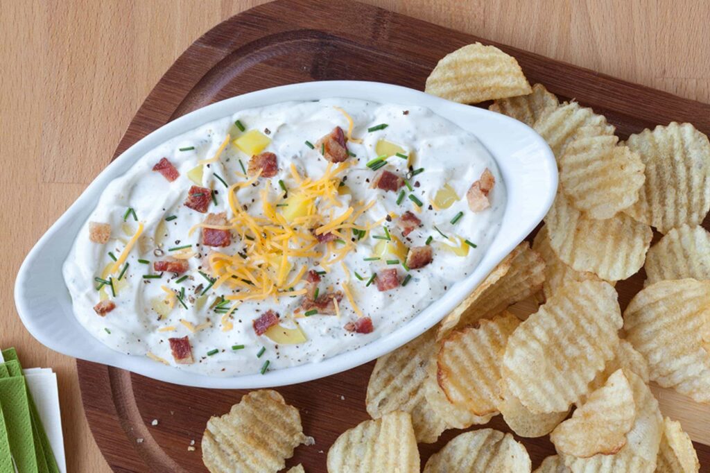 A creamy loaded baked potato dip topped with shredded cheese, bacon, potato pieces, and chives, surrounded by ridged potato chips.