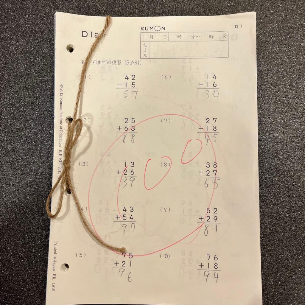 A Kumon worksheet page with ten two-digit addition problems, handwritten answers, red grading marks, and a string threaded through binder holes.