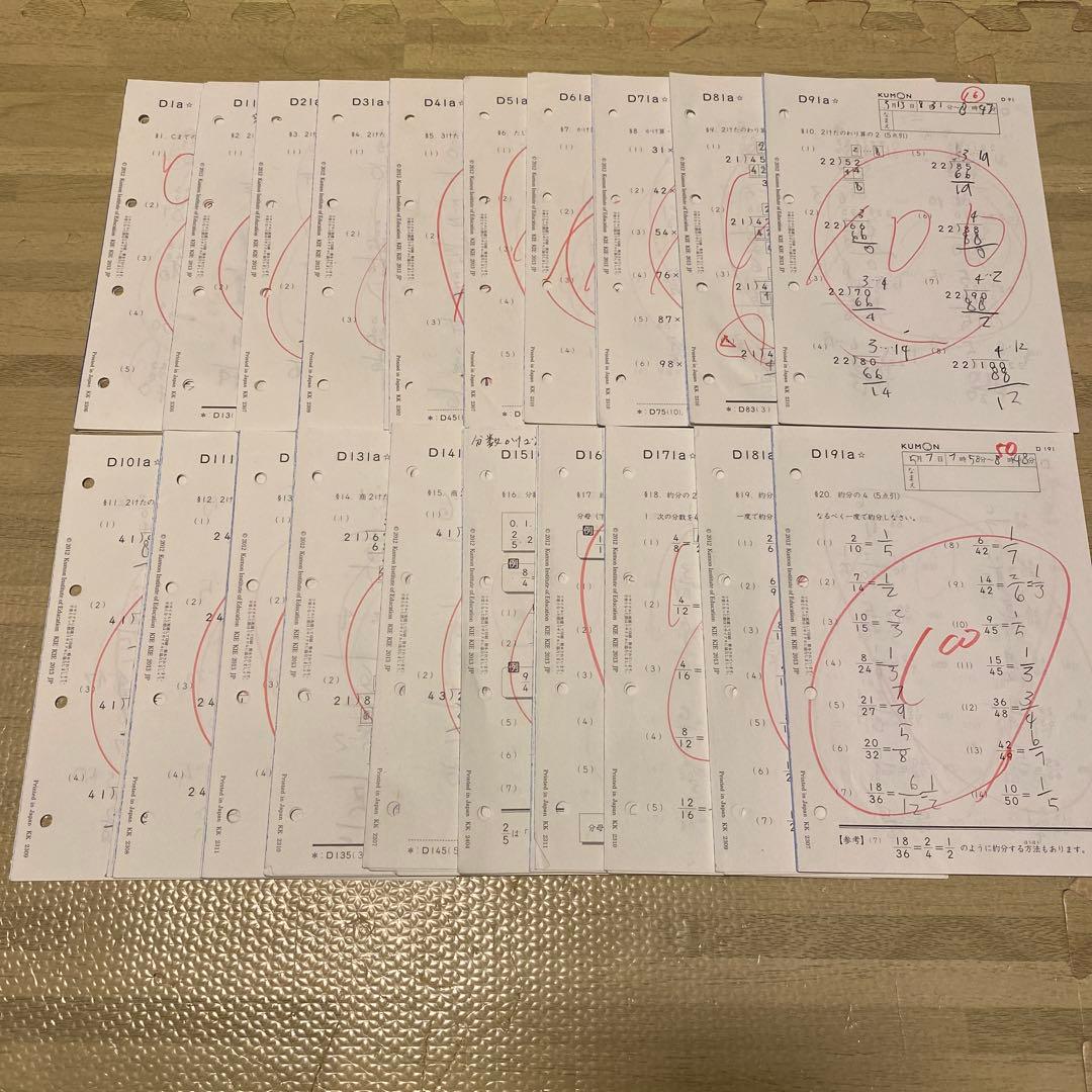 Several Kumon Level D math worksheets are laid out on a floor, showing arithmetic exercises, handwritten answers, and red grading marks.