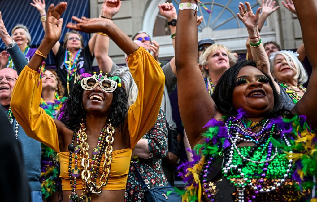 A lively Mardi Gras crowd raises their hands, wearing colorful masks, feathered accessories, and strands of beads.