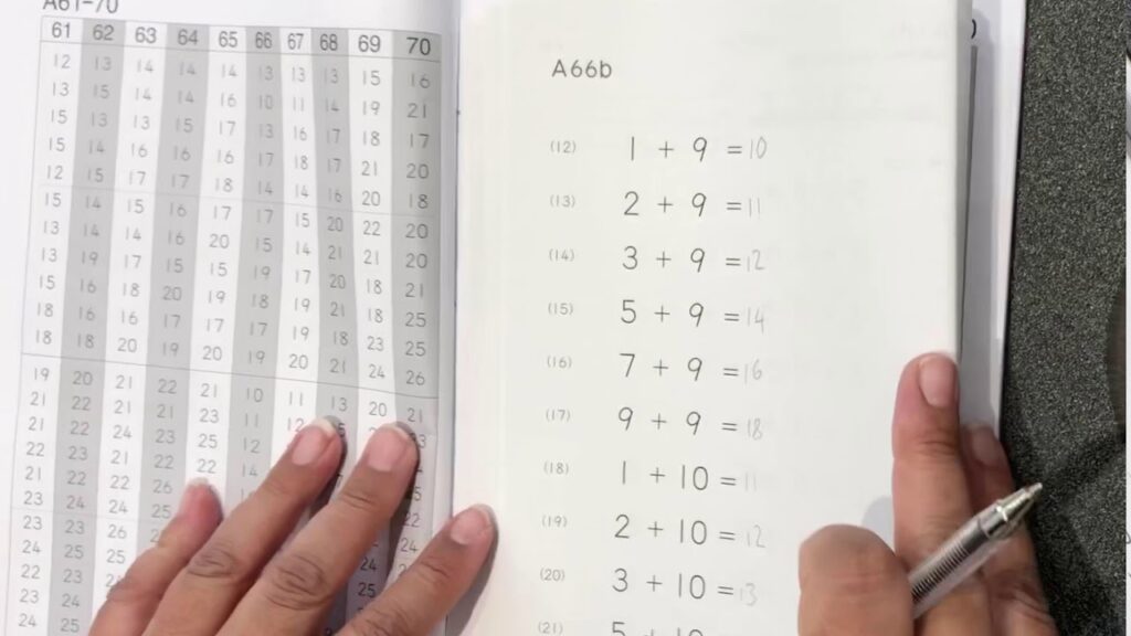 Open math workbook showing an addition exercise page with sums using 9 and 10 beside a numbered grid; hands hold the pages and a pen.