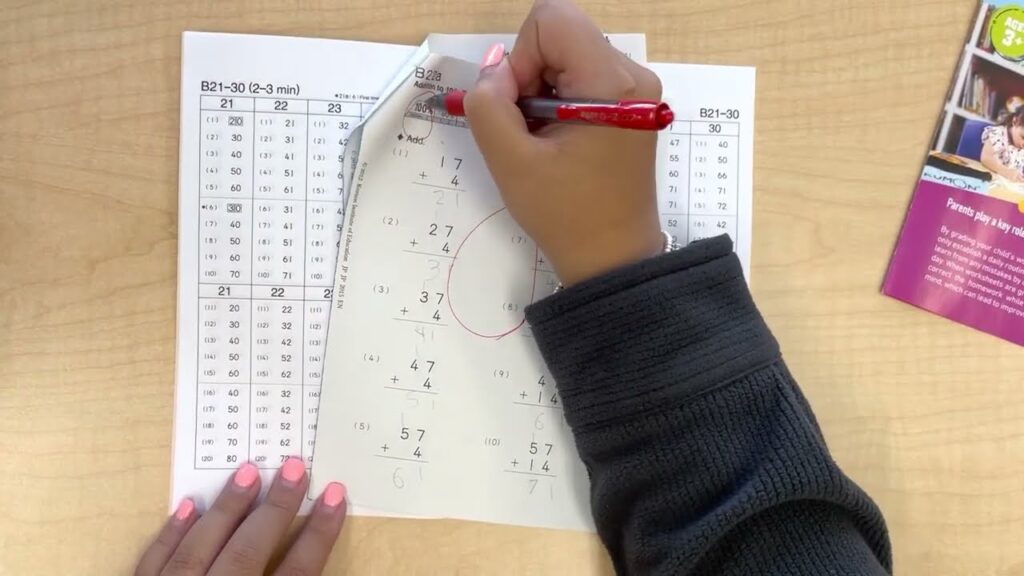 A person writes answers on a Kumon arithmetic worksheet with two-digit addition problems, while other practice sheets lie underneath.