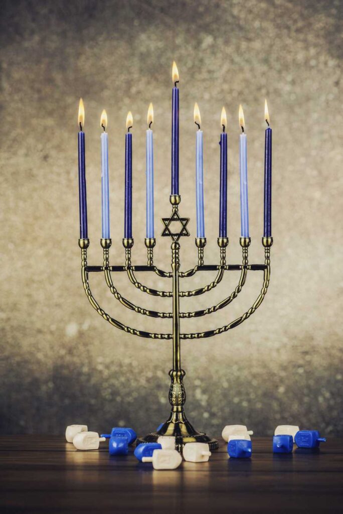 A brass Hanukkah menorah with nine lit candles and a Star of David stands among blue-and-white dreidels on a table.