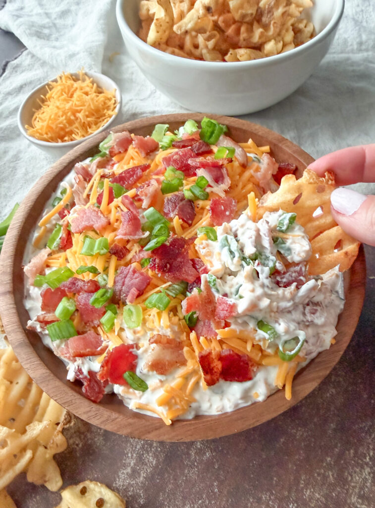 A wooden bowl of creamy dip topped with shredded cheddar, bacon, and green onions, surrounded by potato chips; a hand holds a chip above the bowl.