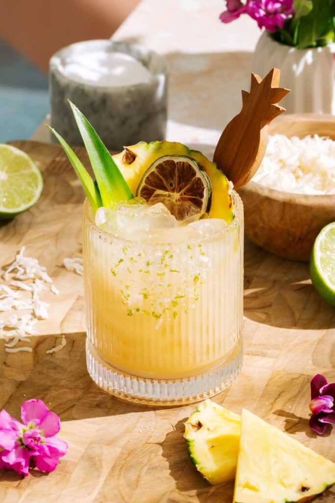 A pale yellow iced drink in a ribbed glass, garnished with lime, pineapple, and green leaves on a wooden board.