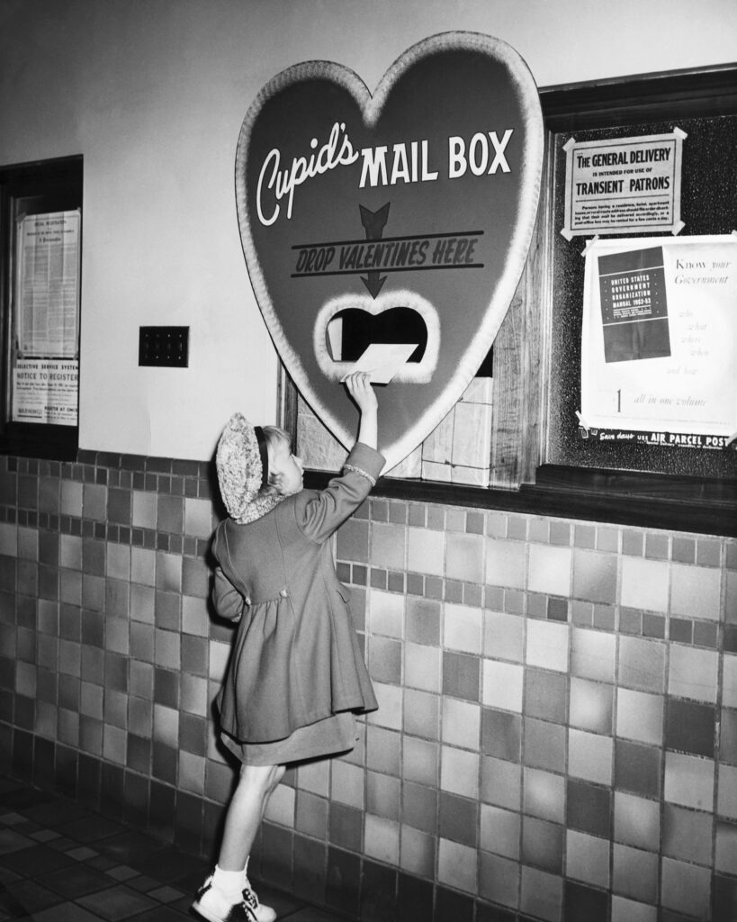 Black-and-white photograph of a child placing a card into a large heart-shaped mailbox labeled “Cupid’s Mail Box” and “Drop Valentines Here.”.