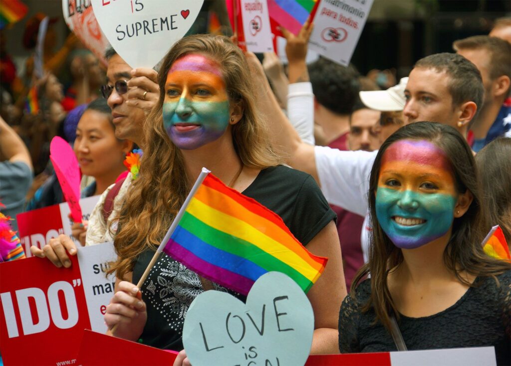 People at a Pride gathering wave rainbow flags; two women in the foreground wear rainbow face paint and hold signs.
